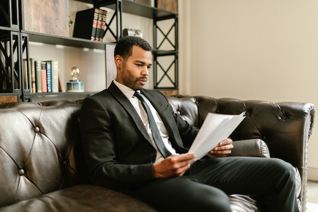 a landlord in a black suit sits on a brown leather couch and reviews a noise-abatement notice before giving it to an individual who caused a noise complaint
