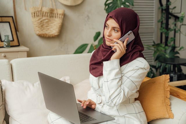 Person wearing a burgundy hijab making a phone call and holding a laptop on their lap
